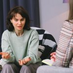 Mulher participa de uma entrevista em uma sala iluminada, diante de outra mulher que a escuta atentamente.