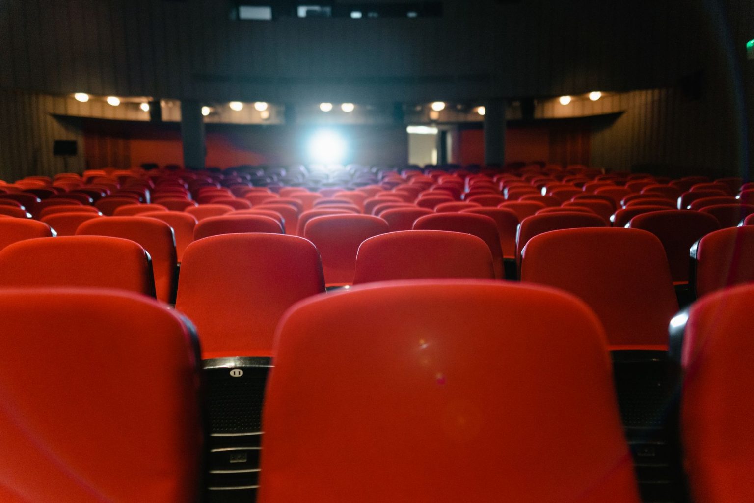 Sala de cinema vazia, com fileiras de poltronas vermelhas voltadas para uma tela iluminada.